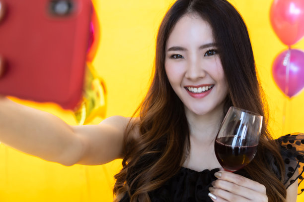 肖像特写的微笑美丽的年轻亚洲女人用手机自拍照片，并拿着一杯与黄色墙壁和颜色满气球派对的红酒。图片下载