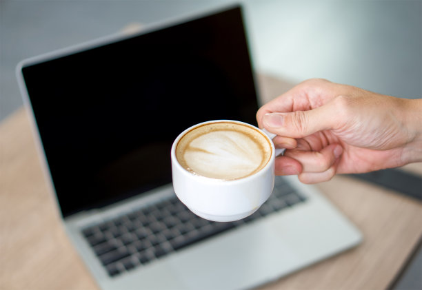 手里拿着一杯拿铁咖啡。背景是黑屏的笔记本电脑。图片下载