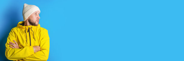 一名身穿黄色夹克的消极年轻人在蓝色背景下双手交叉，向一边看。横幅。图片下载