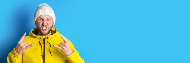 一名身穿黄色夹克、蓄着胡须的年轻人展示了一只摇滚山羊，蓝色背景上有一只角。横幅图片下载