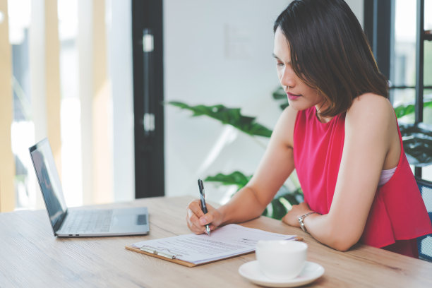 一个快乐的中年亚洲女商人，穿着休闲的服装在家工作，在电脑上检查电子邮件，在财务会计文件纸上写作。库存图片图片下载