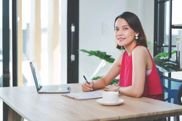 一个快乐的中年亚洲女商人，穿着休闲的服装在家工作，在电脑上检查电子邮件，在财务会计文件纸上写作。库存图片图片下载