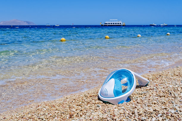 沙滩上的全面罩浮潜面罩，背景是海洋、岛屿、船只和晴朗的天空图片下载