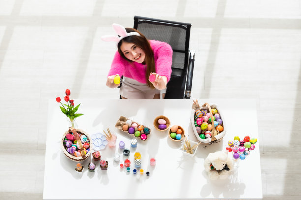 复活节的概念，快乐的亚洲年轻女人戴着兔子耳朵手拿着一个篮子与彩色的复活节蛋在白色木制桌子的背景在顶视图图片下载