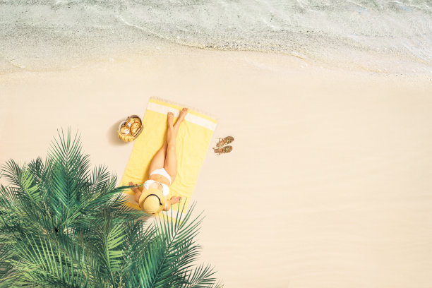 一个戴草帽、穿着比基尼的女人在棕榈树下的黄色沙滩浴巾上晒太阳。暑假女性在沙滩上放松。俯视图图片下载