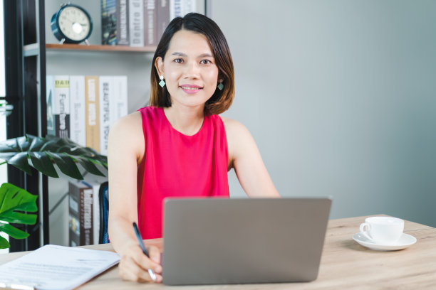 一位快乐的中年亚洲女商人，穿着休闲的休闲装在家工作，在电脑上查看电子邮件，在财务会计凭证上写作。商业库存图片图片下载
