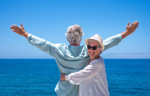 戴着帽子的快乐的老年妇女拥抱着她的丈夫，同时张开双臂看着海面上的地平线。快乐的退休夫妇享受暑假图片下载