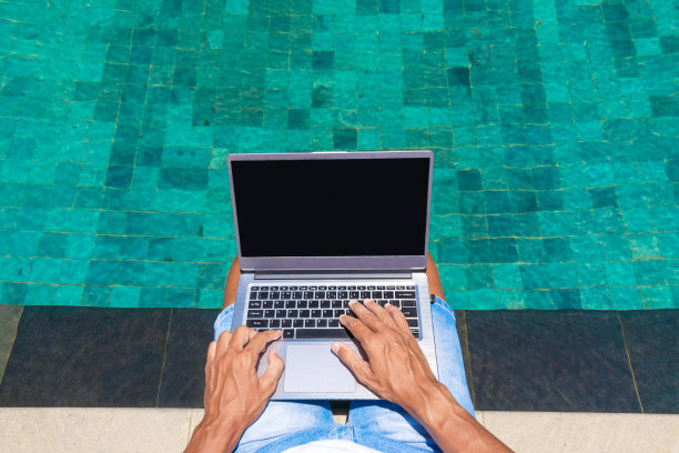 一个男人在游泳池边用笔记本电脑工作。从以上观点。自由职业者，在大自然中工作。手在打字。图片下载