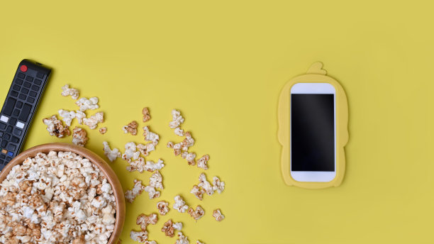 一碗爆米花，黄色背景的遥控器和智能手机。图片下载