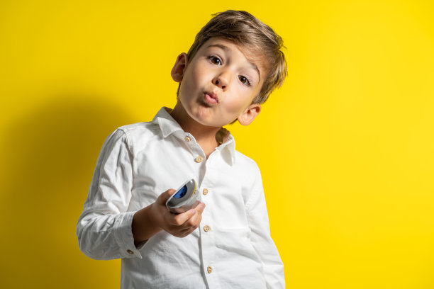 四岁的白人小男孩穿着白色衬衫站在黄色背景前拿着遥控器摆姿势，自信地看着镜头图片下载