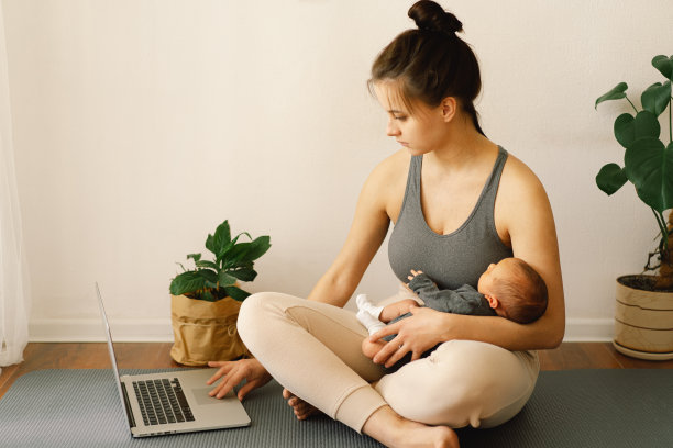 母亲抱着刚出生的儿子，在家里用电脑工作。现代母亲和自由职业。使用技术。图片下载