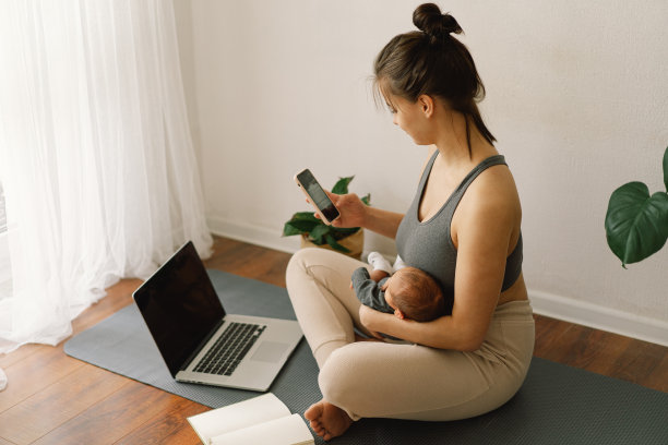 母亲抱着刚出生的儿子，在家里用电脑工作。现代母亲和自由职业。使用技术。图片下载