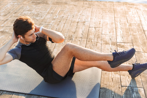 照片上是20多岁的年轻人穿着运动服躺在垫子上，做交叉仰卧起坐，早上在海边的木制码头上锻炼图片下载