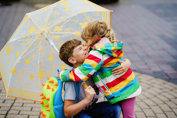 学龄男孩从幼儿园接小妹妹、学前班女孩。两个快乐的孩子撑着伞在雨中玩耍，在坏天气下雨天玩耍。兄弟姐妹的爱。图片下载