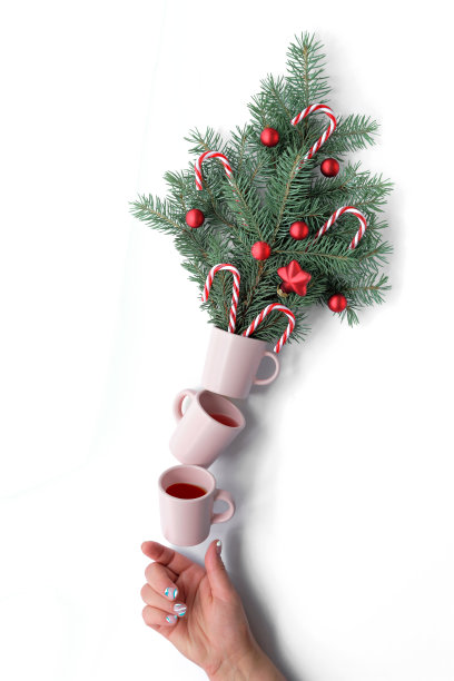 创造性的圣诞平衡安排。咖啡杯支撑着花盆，花盆上装饰着红色的圣诞装饰物和拐杖糖。手支撑着金字塔。创意的圣诞布置图片下载