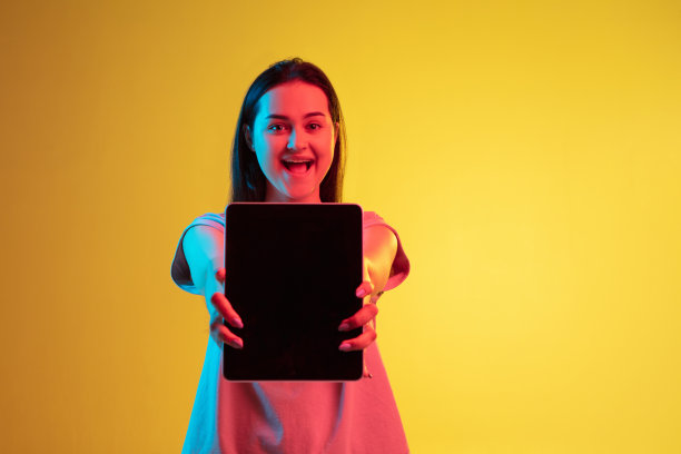 年轻美丽的白人女孩的肖像，在霓虹灯的黄色背景下孤立地拿着写字板。图片下载