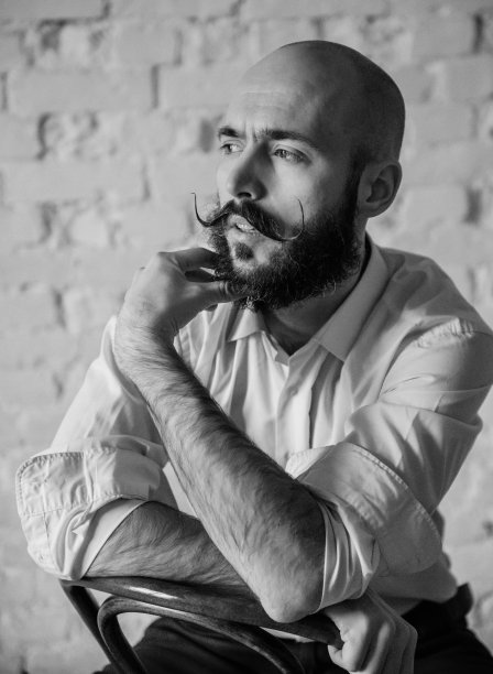 画像中，一个留着胡子和小胡子的年轻秃顶男子穿着一件白色衬衫坐在房间里的椅子上，黑白相间图片下载