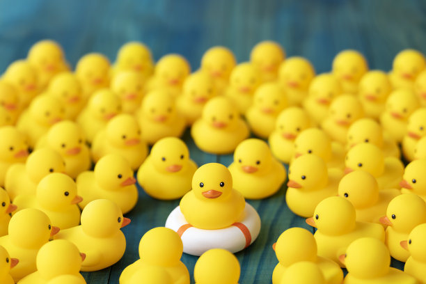 一群黄色橡皮鸭围在一起，看着一只坐在救生圈、救生带或充气圈上的鸭子。场景设置在青绿色的背景上。图片下载