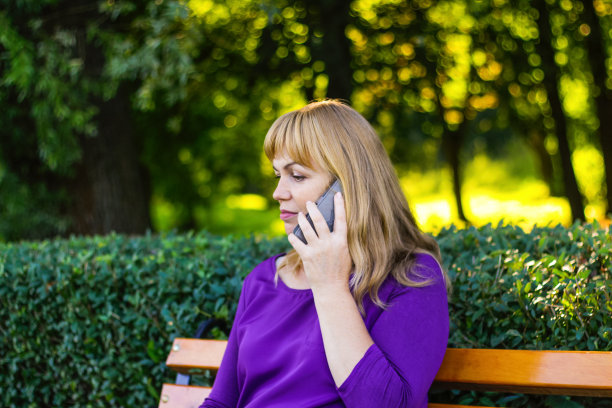 把白种金发女人的镜头移开，在外面打电话，在外面。穿着紫色上衣的40多岁女人在公园的长椅上。成年人使用手机，智能手机。的焦点图片下载