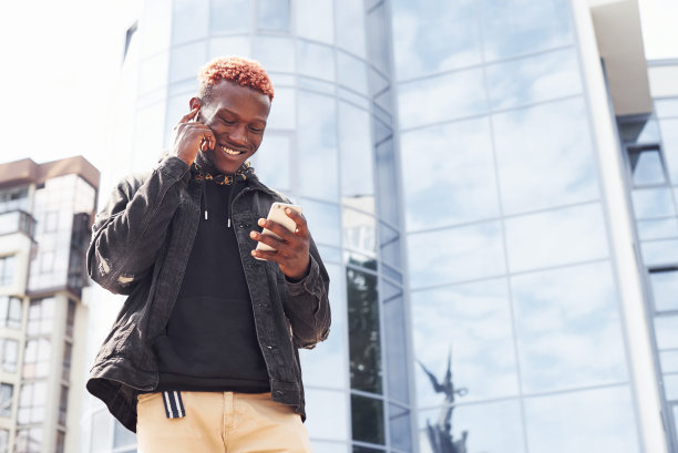 拿着电话。年轻的非洲裔美国人穿着黑色夹克站在城市的户外，对着现代化的商业建筑图片下载