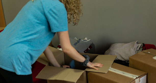 一个漂亮的卷发年轻女人搬进她的新公寓，打开盒子，安排她的衣服和盘子的库存照片。图片下载