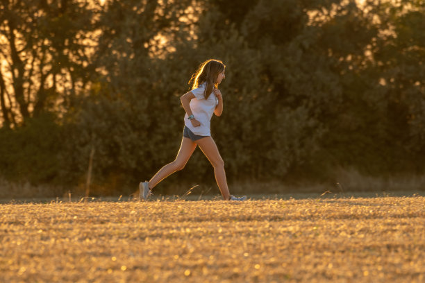 十几岁的女孩，金色长发，短裤和白色t恤跑过收获的田野，背景是树木。图片下载