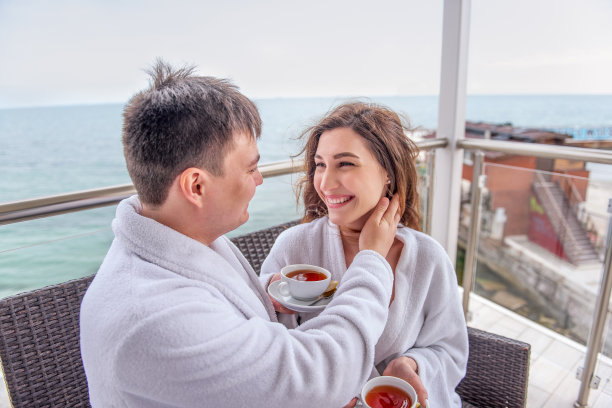 一对穿着白色浴袍的夫妇坐在海边的露天露台上，用杯子喝柠檬茶。情人度假旅行。一个年轻男子为女人拉直头发。新婚夫妇度蜜月的地方图片下载