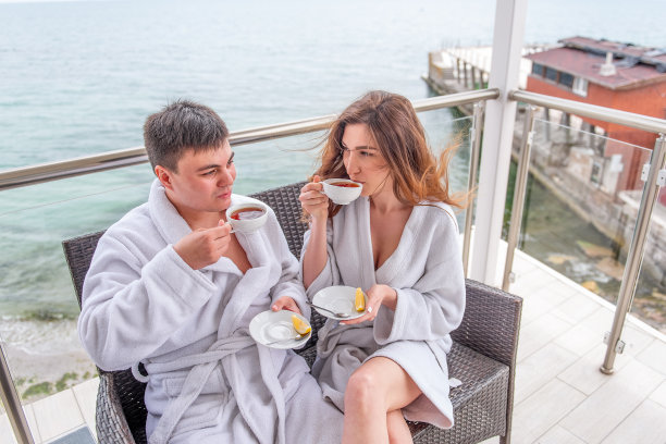 一对穿着白色浴衣的夫妇坐在海边的露天露台上，用杯子喝柠檬茶。情人度假旅行。年轻人和女人拥抱在一起。新婚夫妇在度蜜月图片下载