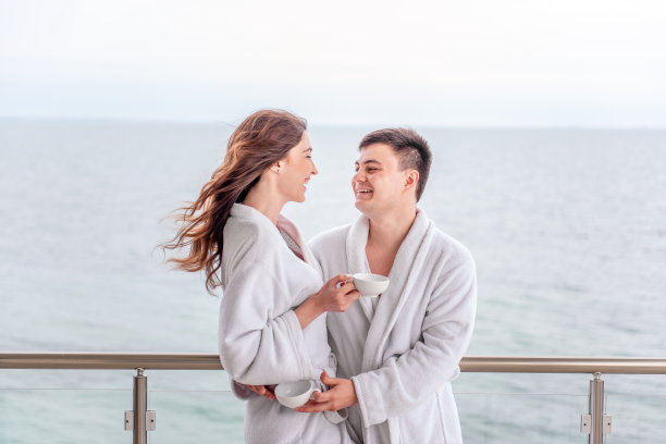 穿着白色毛巾浴袍的年轻情侣在蓝色海景背景下喝茶。新婚夫妇在旅行中度蜜月。相爱的恋人幸福地笑着拥抱，彼此相望，休息。图片下载