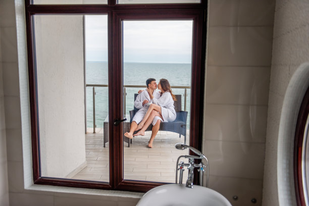 一对穿着白色浴衣的夫妇坐在海边的露天露台上，用杯子喝柠檬茶。情人度假旅行。年轻人和女人拥抱在一起。新婚夫妇在度蜜月图片下载