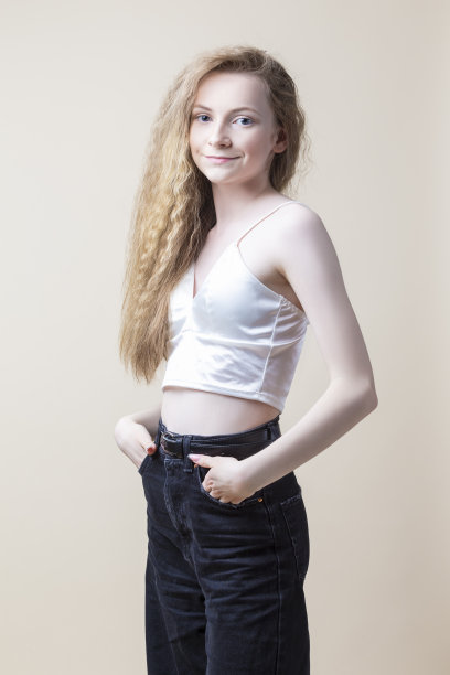 微笑的白人金发女孩穿着白色胸罩与牛仔裤和摆姿势在米色背景。垂直拍摄图片下载