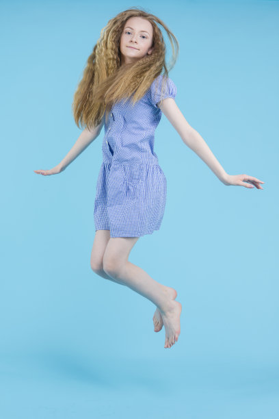 积极快乐跳跃的十几岁的金发女孩在蓝色的背景蓝色长裙。垂直的图像组成图片下载