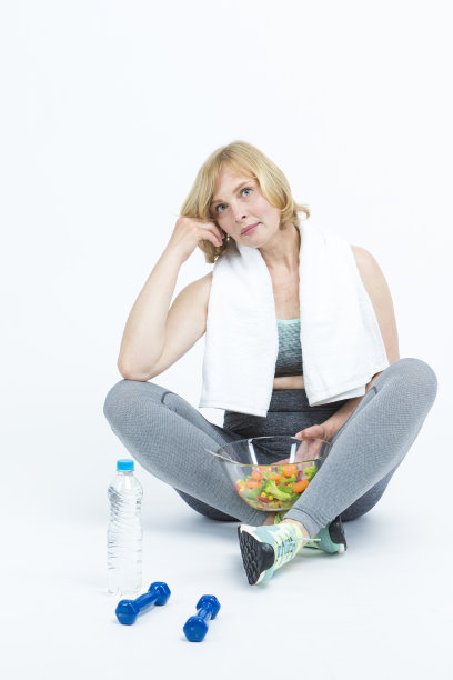 成熟的白人妇女在健身训练在专业运动服与水瓶和碗新鲜蔬菜在白色。垂直拍摄图片下载