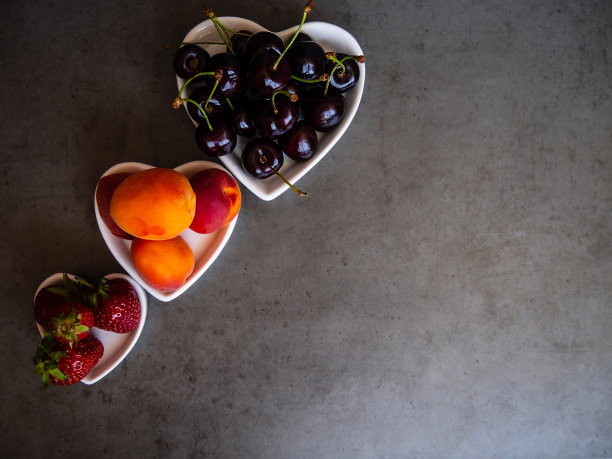 灰色的混凝土桌子上，一个白色的心型盘子里放着新鲜、成熟的草莓、樱桃和杏子。贝瑞。背景和纹理。图片下载