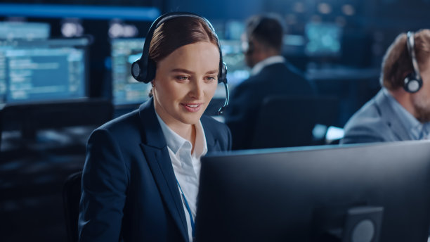 在一个黑暗的监控室里，满是同事和显示屏，快乐美丽的技术客户支持专家一边用耳机说话，一边用电脑工作。图片下载