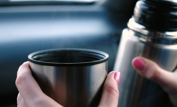 特写的女人的手保持一杯热茶和热水瓶坐在汽车在冬天。图片下载