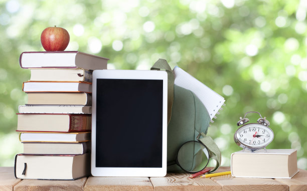 木桌上放着一堆书，里面有红苹果、银钟、数码板、彩色铅笔、放大镜、白色笔记本和书包图片下载
