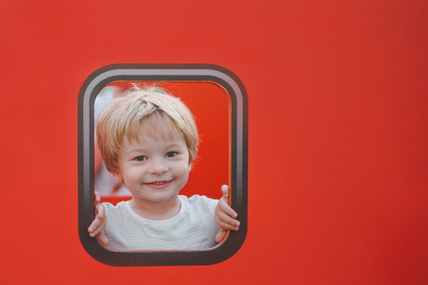 一个美丽的微笑的孩子的肖像窥视玩具纸板在一个夏天的操场上的红色窗户图片下载