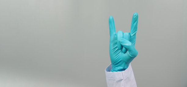 手是做魔鬼角手势，穿着医生袍和蓝色医疗手套在灰色的背景。工作室拍摄。图片下载