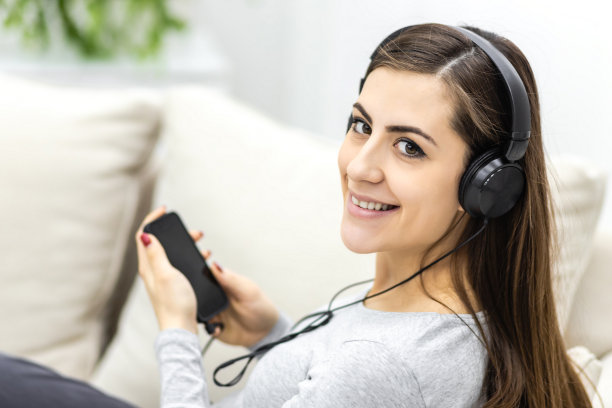 孕妇坐在沙发上用耳机听音乐和使用手机的照片。图片下载