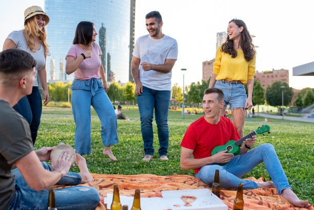 年轻的男孩们弹奏着尤克里里和铃鼓，人们在草坪上赤脚跳舞，PIC尼克与城市轮廓的场景，千禧一代在啤酒和披萨聚会，夫妇在公园里玩耍图片下载