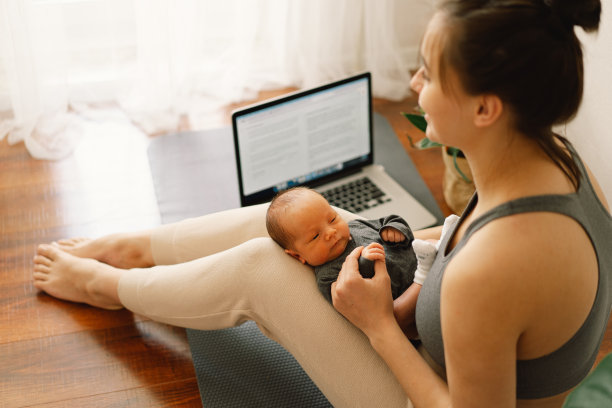 母亲抱着刚出生的儿子，在家里用电脑工作。现代母亲和自由职业。使用技术。图片下载