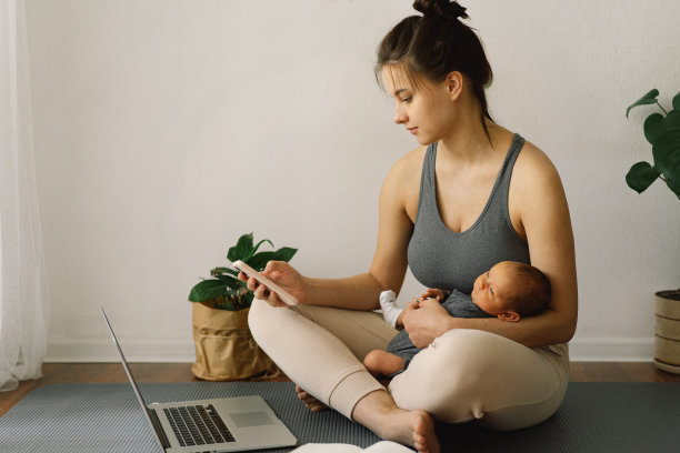 母亲抱着刚出生的儿子，在家里用电脑工作。现代母亲和自由职业。使用技术。图片下载