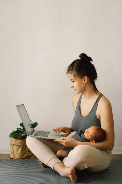 母亲抱着刚出生的儿子，在家里用电脑工作。现代母亲和自由职业。使用技术。图片下载