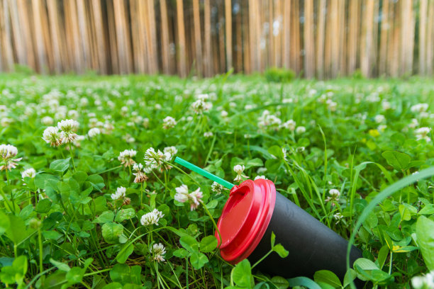 一个黑色的纸咖啡杯和一根吸管被扔到绿色的草地上。生活垃圾对环境的污染。题字俄文:小心，烫手图片下载