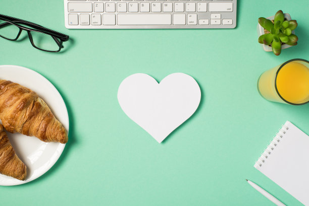 俯视图照片的工作场所键盘，花盆眼镜提醒笔盘与羊角面包杯果汁和白纸心在中间孤立的绿松石背景与空白图片下载