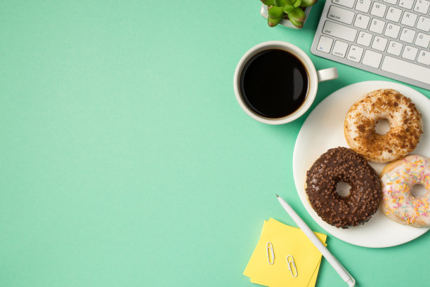 俯视图照片的工作场所键盘，笔夹，便利贴，纸，花盆，杯饮料和三个甜甜圈在盘子上孤立的绿松石背景与copyspace图片下载
