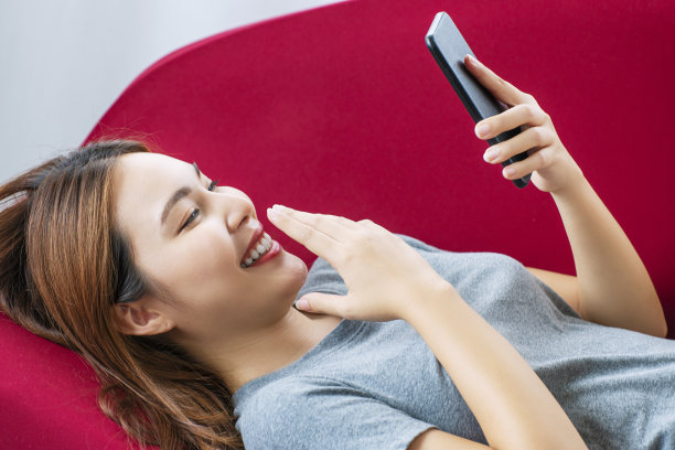 生活方式的技术概念。欢快的年轻女子拿着智能手机，躺在客厅的红色沙发上休息。随便的女孩在社交媒体上用手机发信息。图片下载