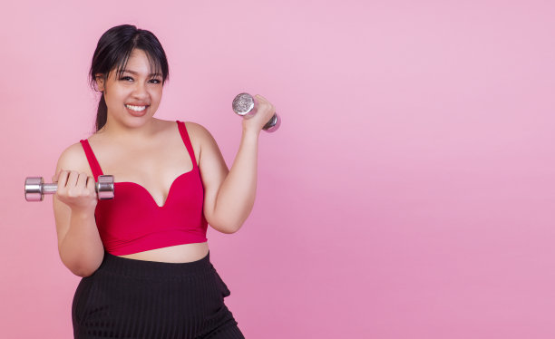 运动健身饮食保健理念。胖嘟嘟的胖女人微笑着举着哑铃减肥，而站在孤立的粉色背景。图片下载
