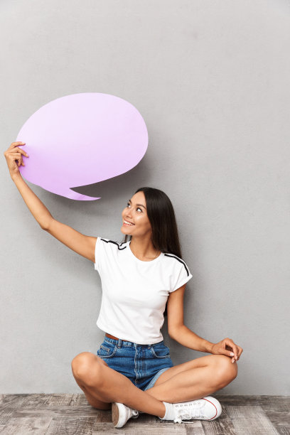 快乐的女人穿着休闲服装拿着空白的气球与copyspace，而坐在地板上双腿交叉孤立的灰色背景图片下载